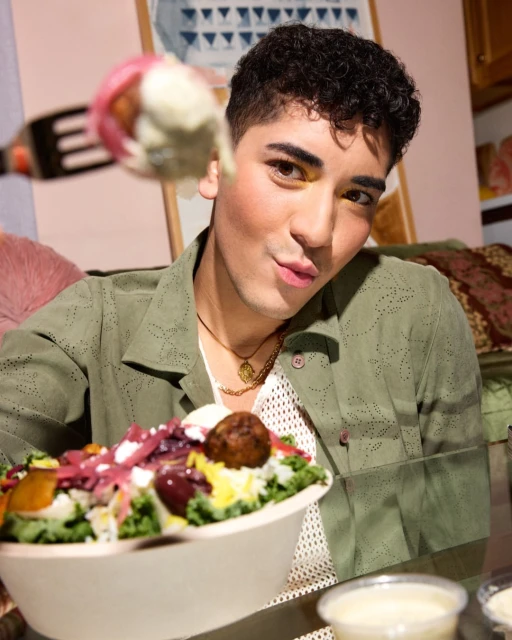 Person eating a salad with a fork. Close-up of a person with curly dark hair, light green shirt, and a white bowl of salad.