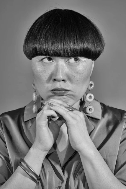 Dramatic black-and-white studio portrait of an Asian woman with a bowl cut, striking makeup, jewelry, and a reflective blouse.