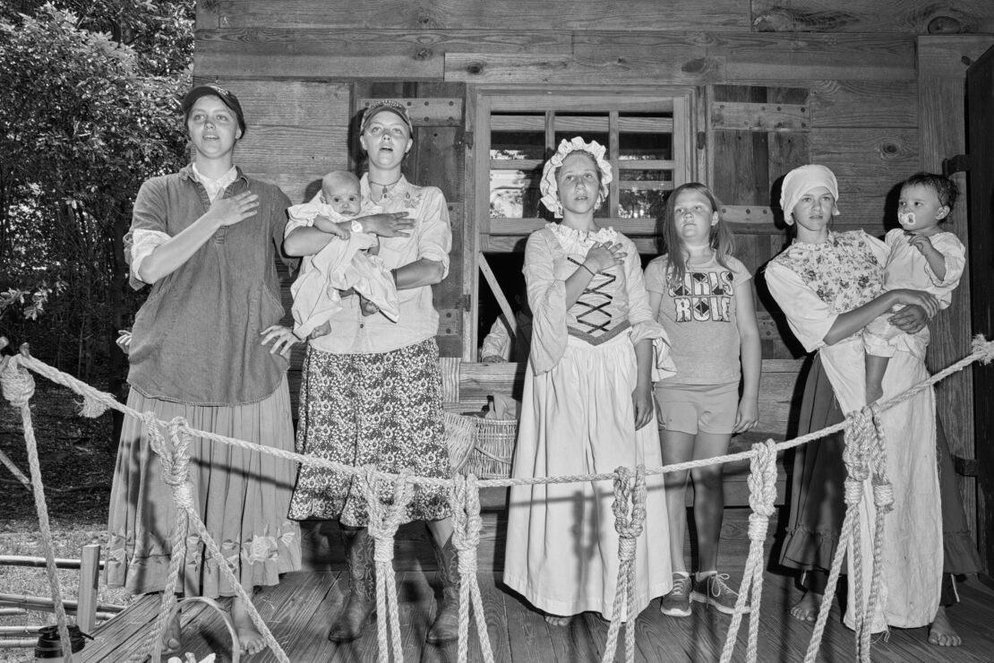 Group of women and children stand in front of a wood building, dressed in vintage and modern clothing. Black and white photo.