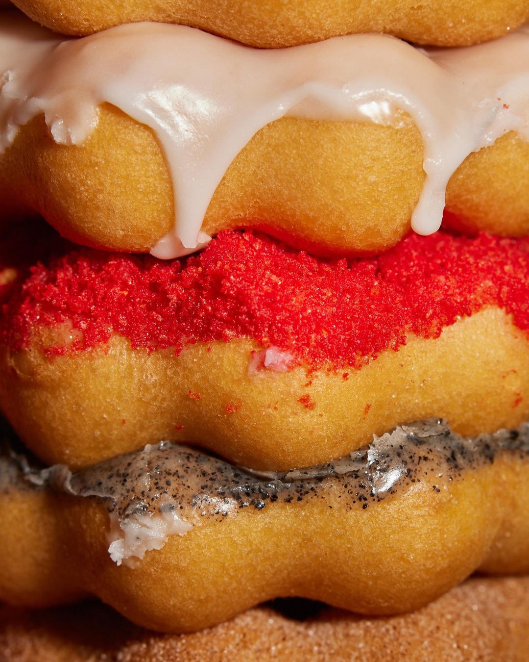 Close-up of a stacked doughnut with various toppings, including glaze, red sprinkles, and a grey speckled coating. Food photography.
