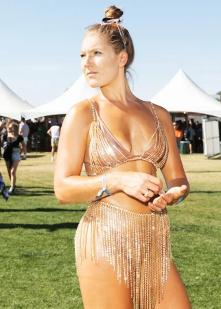 Woman poses in gold chain bikini and skirt at outdoor festival with tents and a crowd in the background on a sunny day.