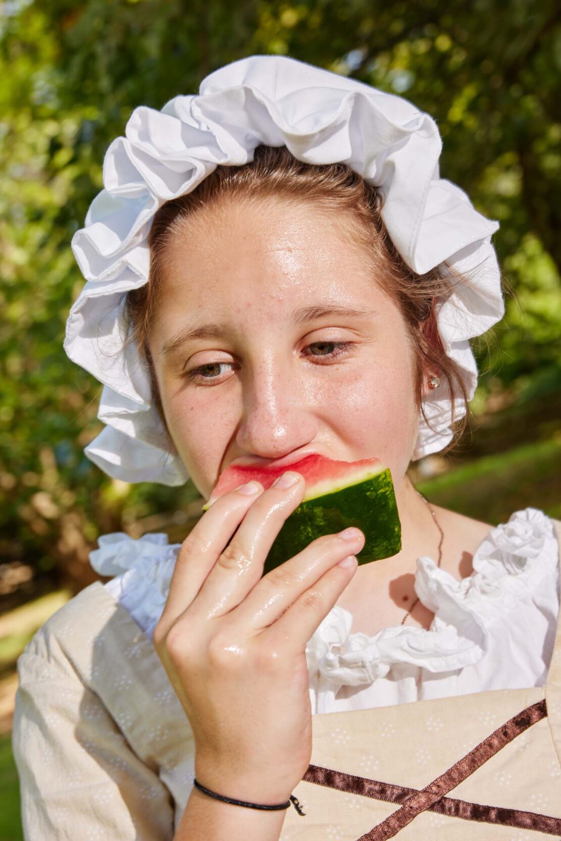 Close-up of a smiling young woman with a ruffled bonnet, enjoying a slice of watermelon outdoors on a sunny day.