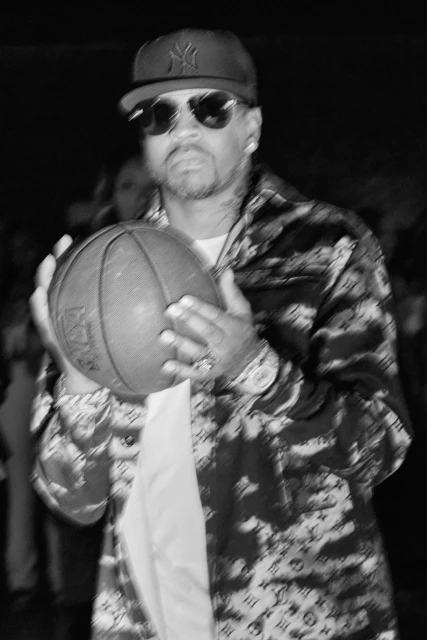 Allen Iverson in black and white photo holding a basketball, wearing sunglasses and baseball cap indoors with people in background.