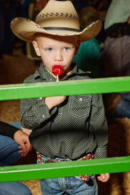 Young boy in a cowboy hat enjoys a red lollipop while standing in front of a green metal railing indoors, wearing a patterned shirt.