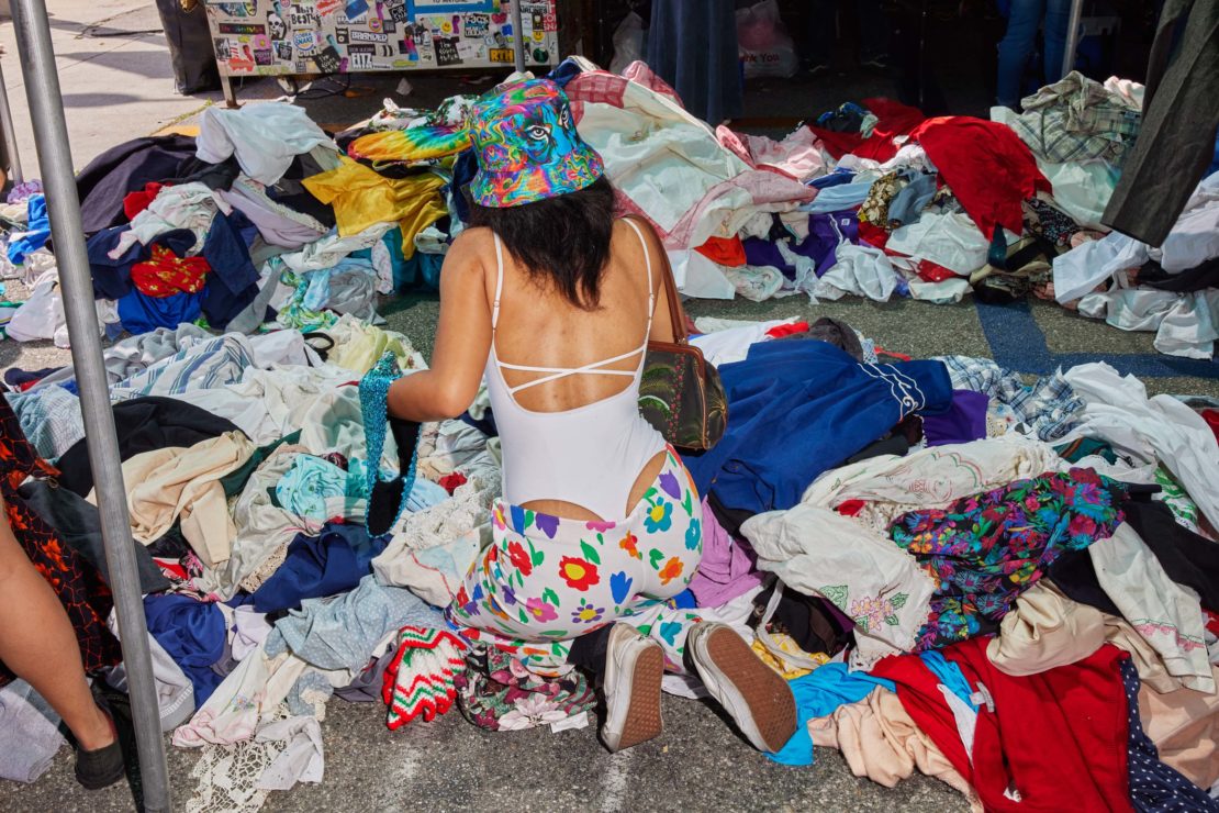 Woman in white swimsuit and floral pants searches through a large pile of clothes at an outdoor flea market on a sunny day.