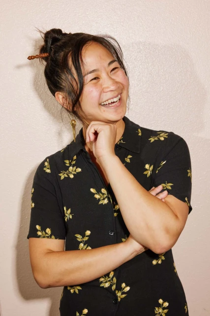 Asian woman in floral dress smiles with hand to chin. Updo hairstyle with chopstick. Portrait on light neutral backdrop, eye-level shot.