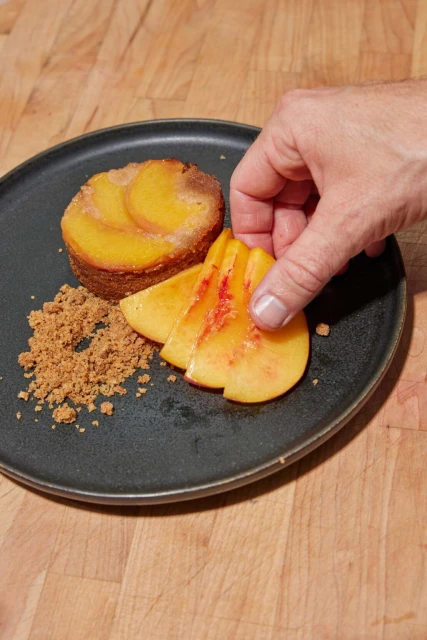 Peach cake with sliced peaches and crumble on a black plate being garnished with a human hand, sitting on a wooden surface.