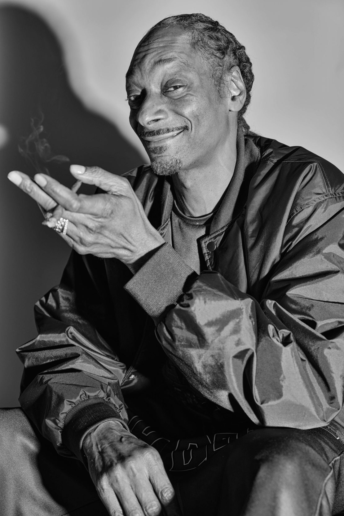 Monochrome portrait of Snoop Dogg smiling and holding up a hand with a ring against a plain studio background.