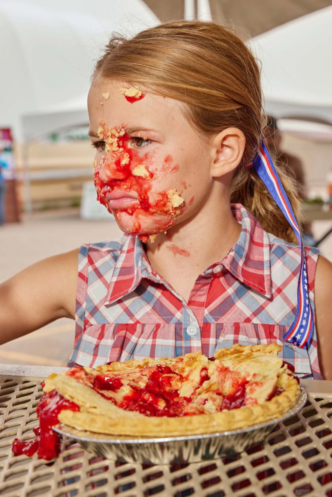 Young girl with pie filling smeared on face, wearing a red and blue plaid shirt, sitting at a picnic table with a cherry pie.