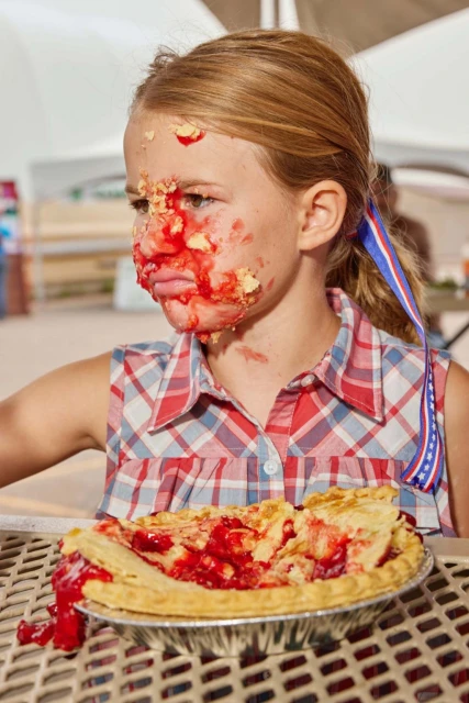 Young girl with pie filling smeared on face, wearing a red and blue plaid shirt, sitting at a picnic table with a cherry pie.