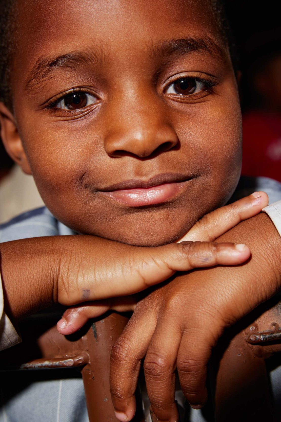 Close-up portrait of a young Black child with brown eyes, resting their chin on their hands, against a dark, softly blurred background.