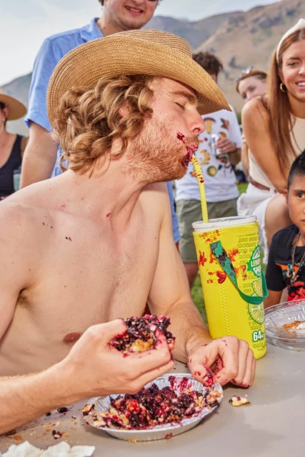 A shirtless man in a cowboy hat enthusiastically eats pie with a crowd in the background on a sunny day, creating a candid, outdoor scene.