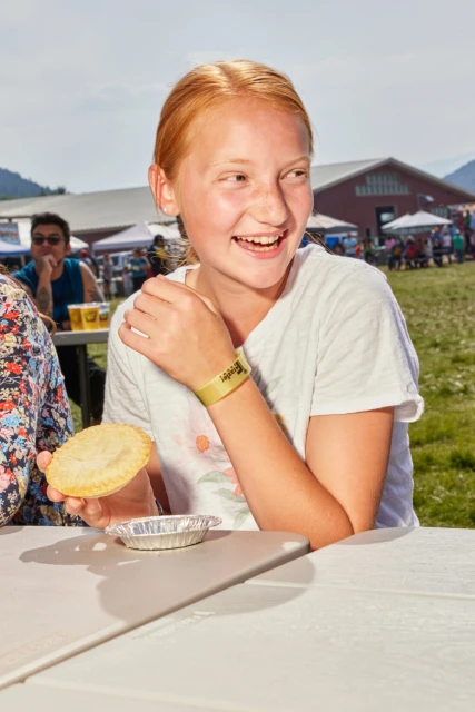 Smiling red-haired child seated at a table with pie at an outdoor event with crowds, tents, and a red barn in the background.