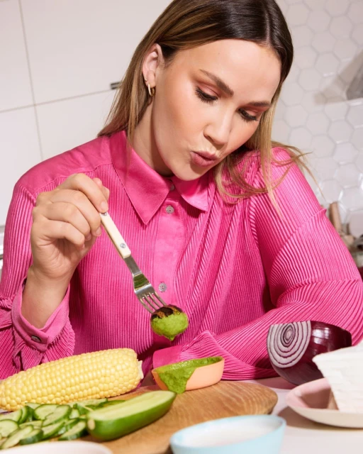 Woman in pink eating green ball with fork at kitchen table with corn, cucumber, and sliced red onion. Healthy eating concept.
