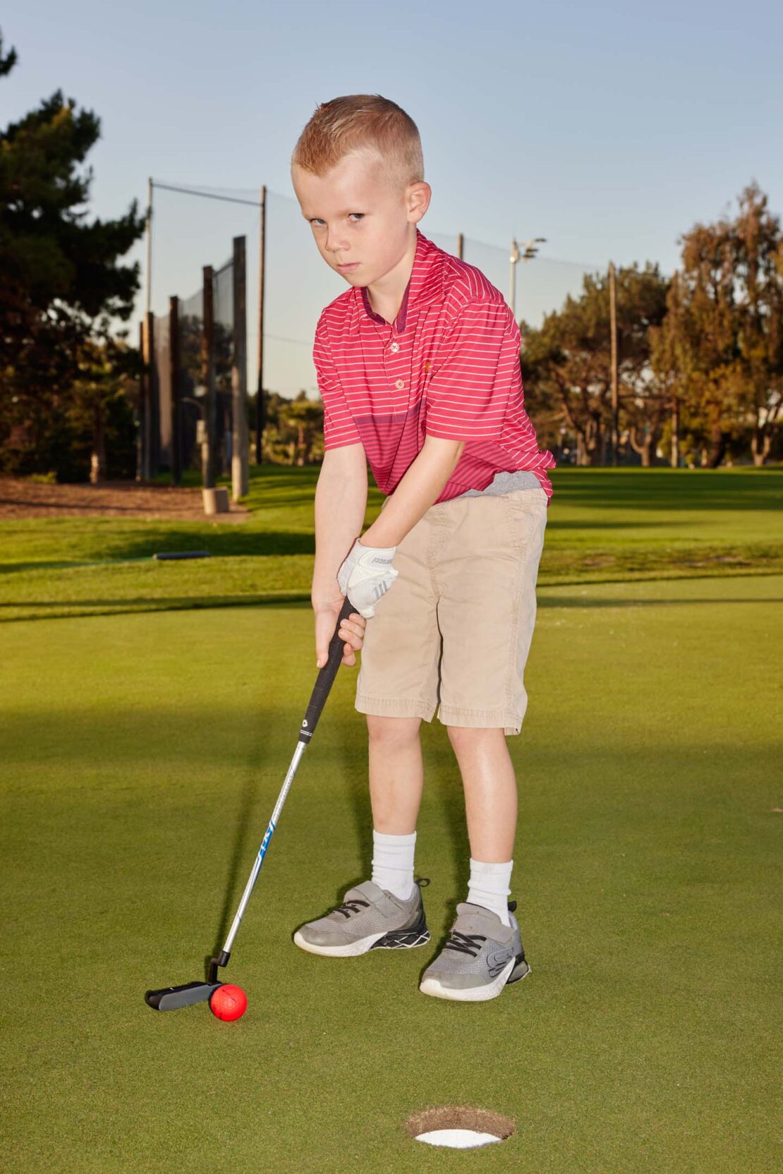 Young boy practices golf putting on a green course with a golf club and red ball, getting ready to swing toward the hole.