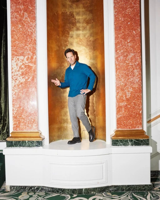 A man in a blue sweater and grey pants stands in a white alcove with marble columns and a gold background, looking at the camera.