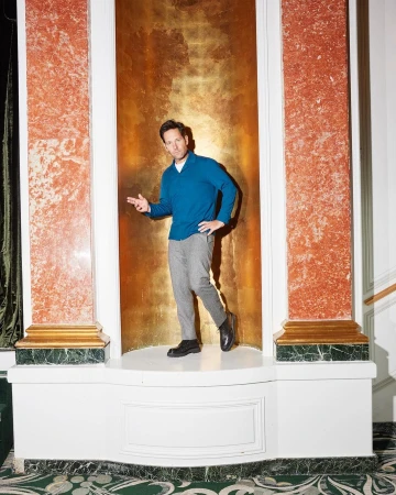 A man in a blue sweater and grey pants stands in a white alcove with marble columns and a gold background, looking at the camera.