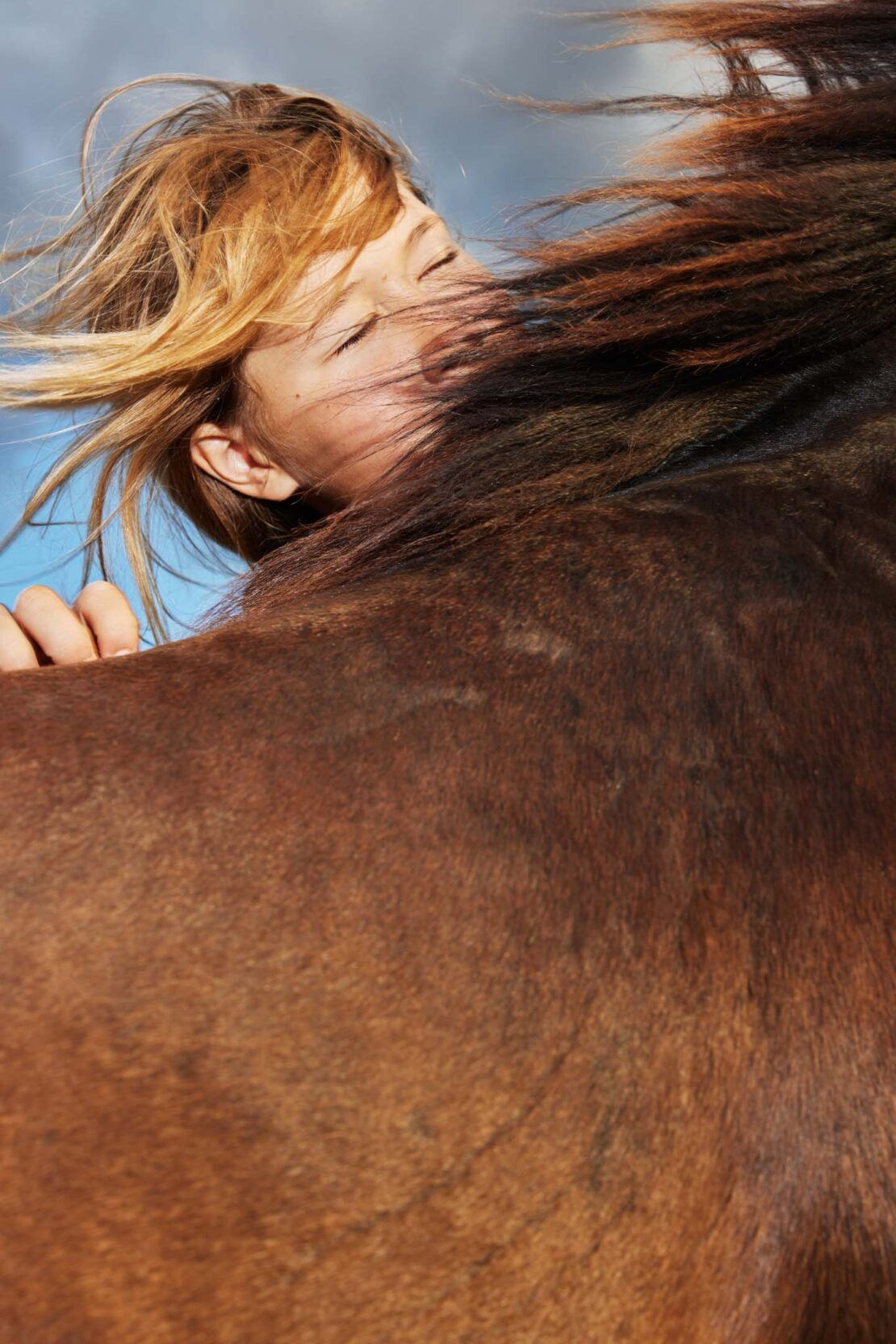 Close-up portrait of a child with eyes closed resting against a brown horse under a blue sky
