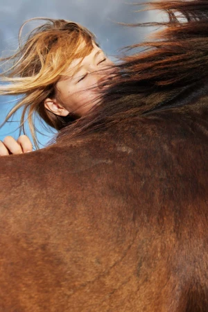 Close-up portrait of a child with eyes closed resting against a brown horse under a blue sky