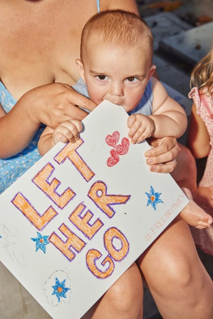 Close-up shot of a baby sitting on an adult's lap, holding a handwritten sign that says 'LET HER GO,' adorned with hearts and stars.