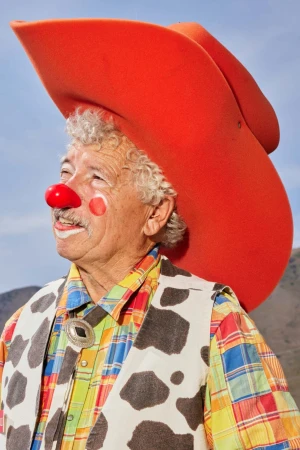 Elderly man wearing an orange cowboy hat, clown makeup, and a cow-print vest, outdoors against a blue sky and mountain background.