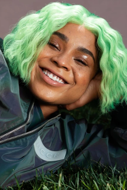 Smiling woman with green hair wearing iridescent jacket on green grass, portrait, studio lighting and background.