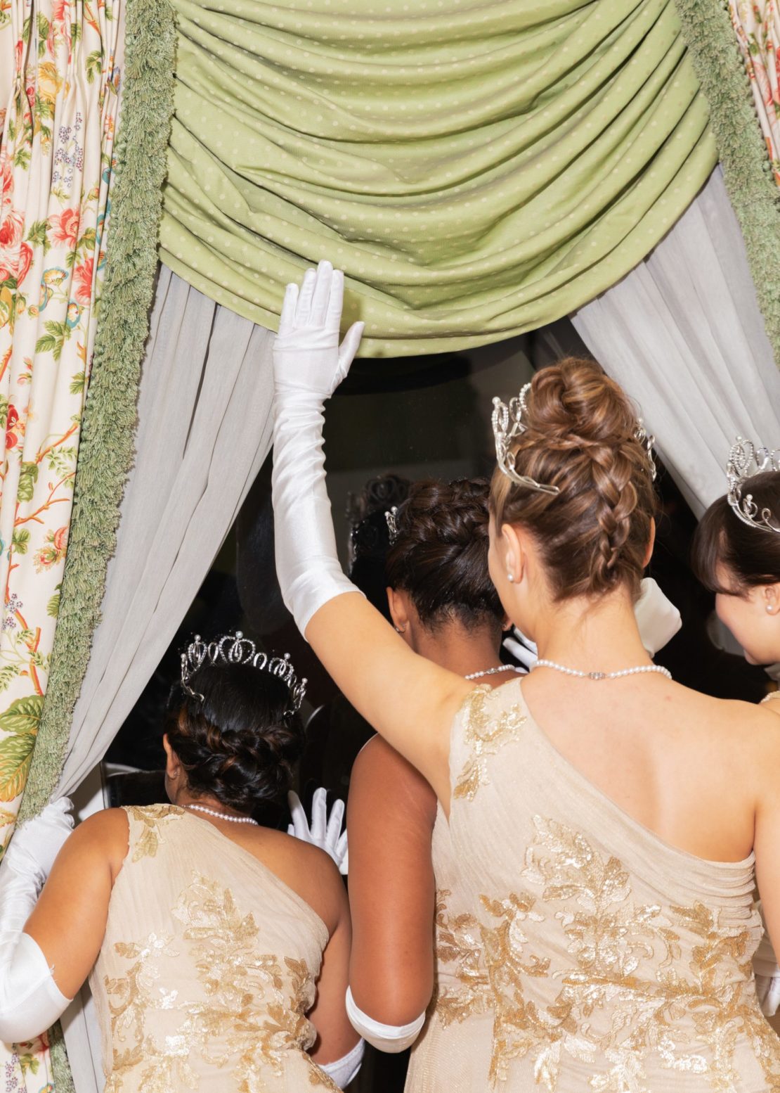 A group of women in formal gowns and tiaras stand near a window, with one woman raising her gloved hand toward the curtain.