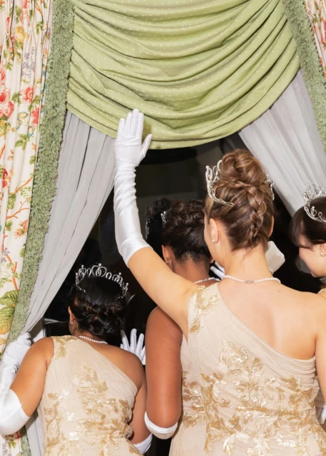 A group of women in formal gowns and tiaras stand near a window, with one woman raising her gloved hand toward the curtain.
