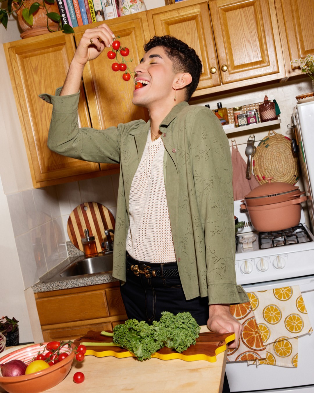 Young adult eating cherry tomatoes in a sunlit kitchen. He's wearing a green jacket over a mesh top in an indoor cooking setting.