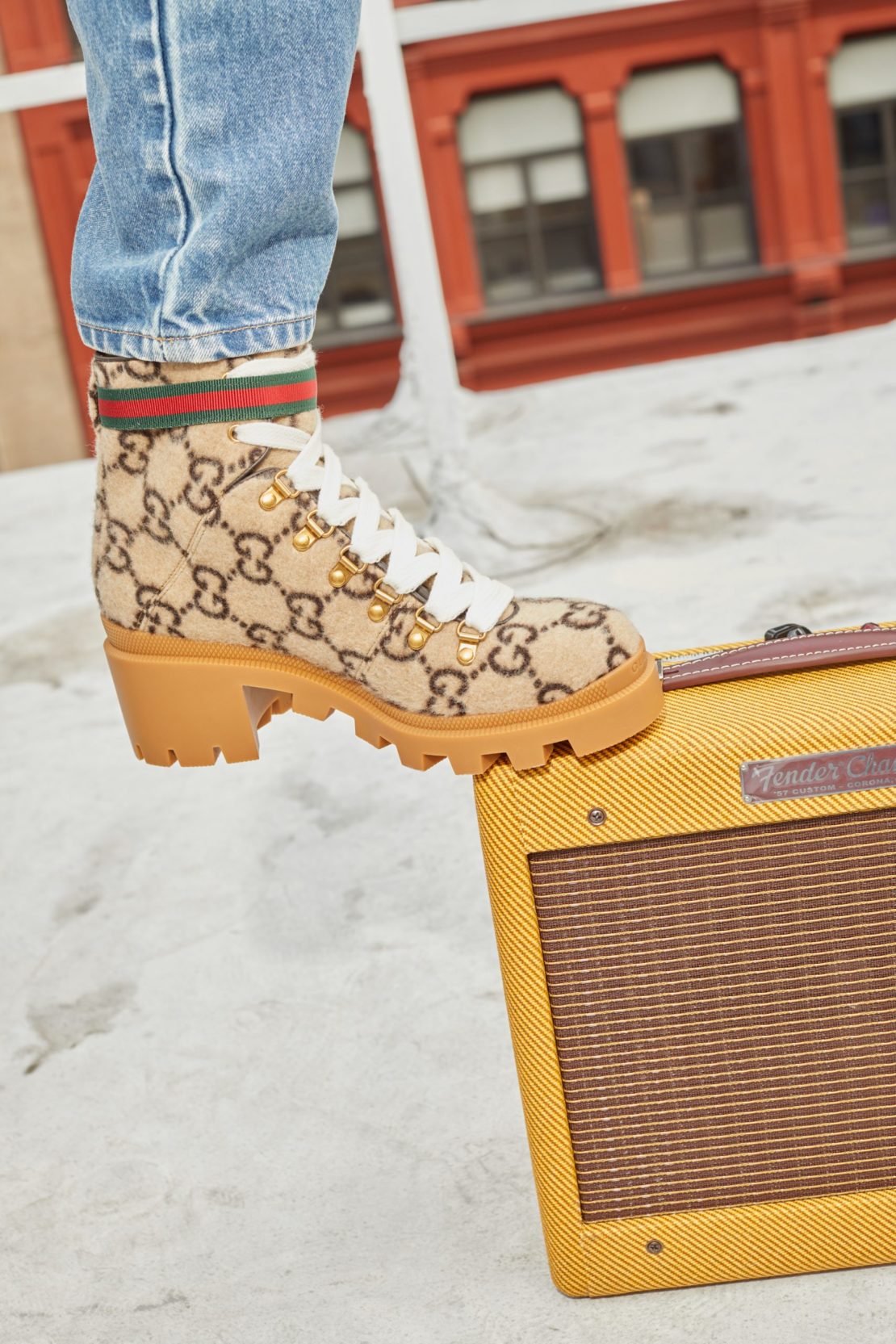 A Gucci ankle boot with a chunky heel is placed on top of a vintage yellow Fender amplifier against an outdoor cityscape backdrop.