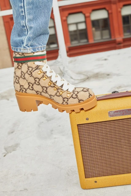 A Gucci ankle boot with a chunky heel is placed on top of a vintage yellow Fender amplifier against an outdoor cityscape backdrop.