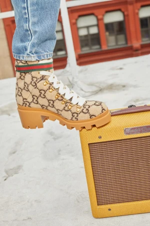 A Gucci ankle boot with a chunky heel is placed on top of a vintage yellow Fender amplifier against an outdoor cityscape backdrop.