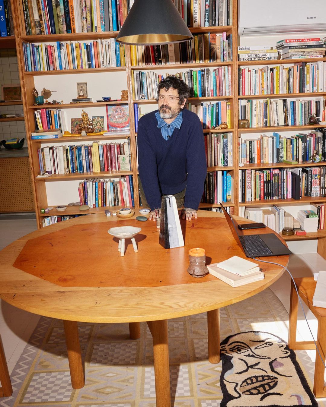 Adult male with beard and blue sweater stands behind round wooden table in a home library with floor rug and a lamp above.