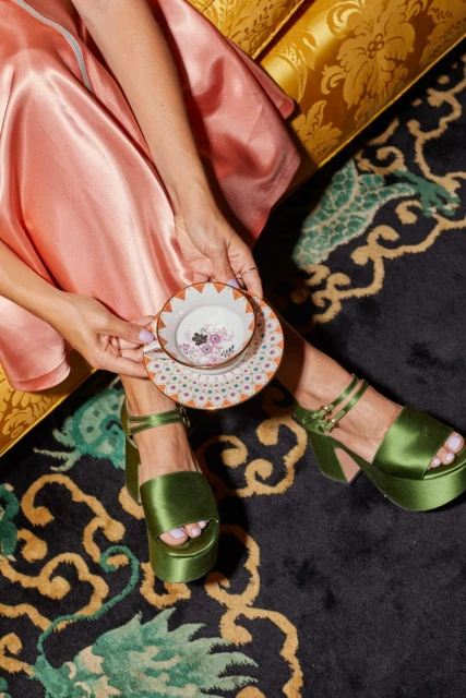 Overhead shot of woman in silk dress and green platform heels holding a teacup and saucer over patterned rug and damask couch.