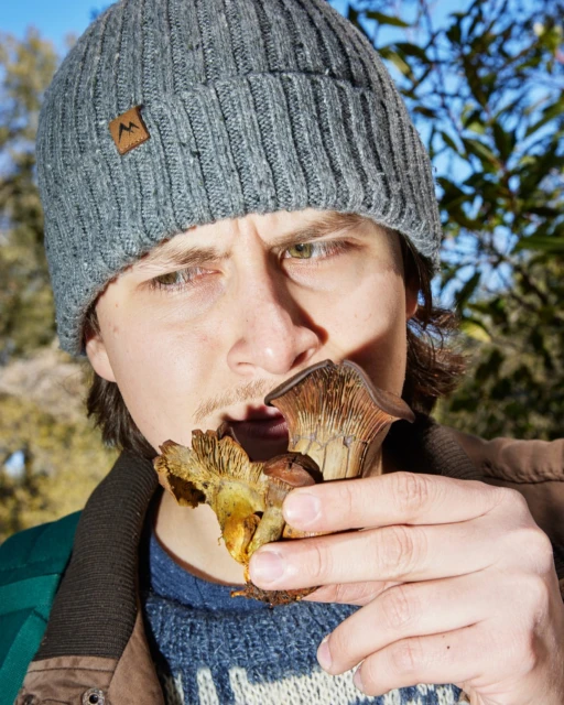 Adult male outdoors in natural light wearing knit hat holding a mushroom near his mouth, with blurred trees and sky background.