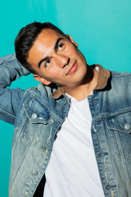 Portrait of a young adult man with dark hair in a denim jacket, looking up against a vibrant aqua studio backdrop.