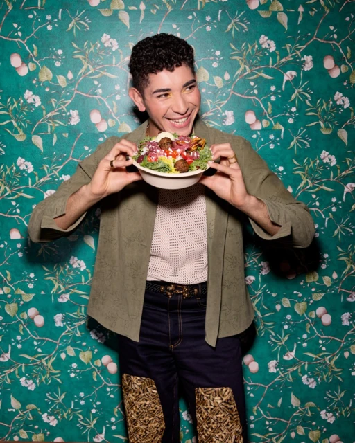 Smiling man in olive jacket holds salad bowl against floral wallpaper. He is wearing dark denim pants and tan patterned boots.