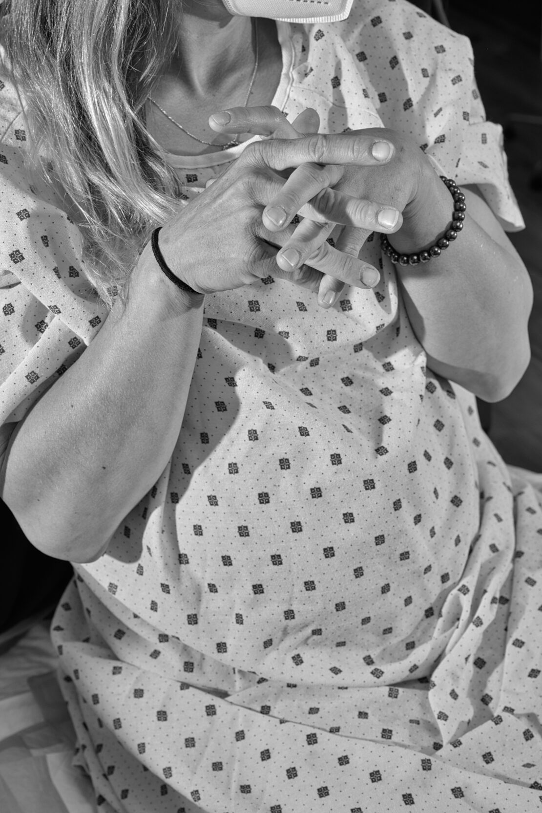 Black and white shot of a fair-skinned adult female patient in a patterned hospital gown, sitting with her fingers interlaced.