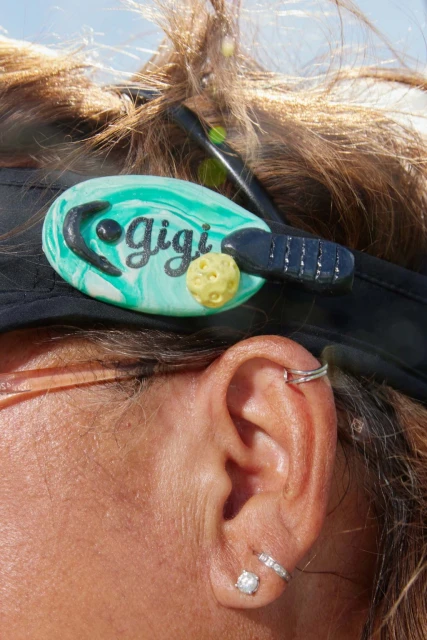 Woman's ear and hair, wearing earring jewelry, and a decorative hairpin of a pickleball racket and ball on a hat.