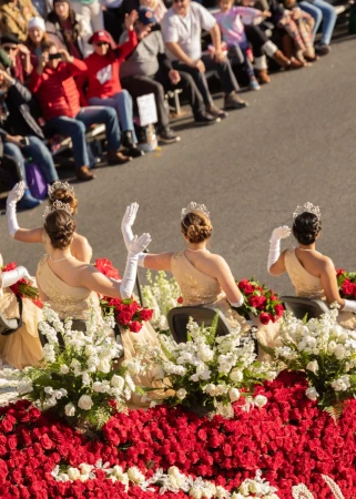 Four women wearing tiaras and evening gowns waving from a parade float decorated with red and white roses, photographed by Maggie Shannon.