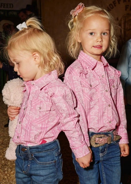 Two young blonde girls in matching pink paisley shirts and blue jeans hold hands indoors, one carrying a teddy bear.