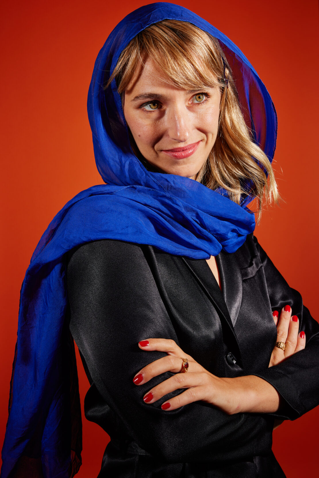 Close-up studio portrait of a smiling adult woman with a blue headscarf and black satin top against a solid orange backdrop.