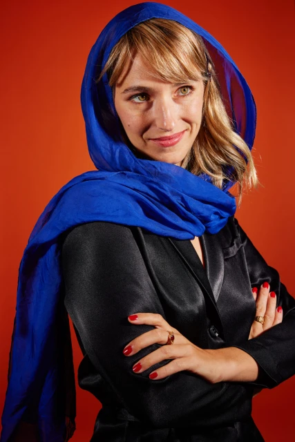 Close-up studio portrait of a smiling adult woman with a blue headscarf and black satin top against a solid orange backdrop.