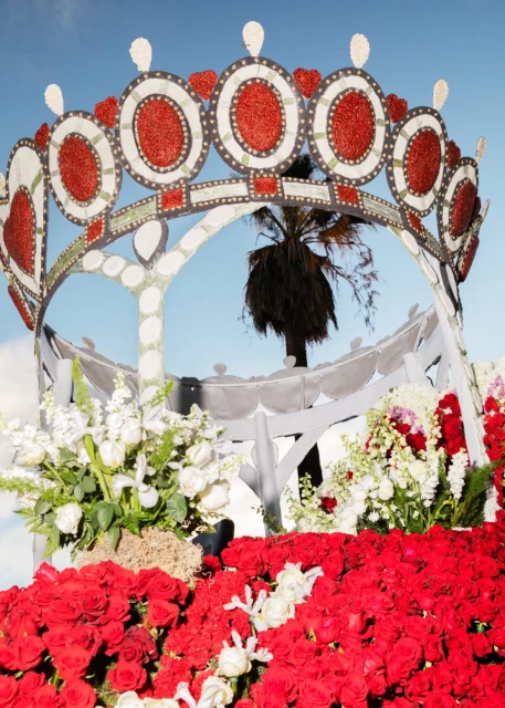 Rose Parade float with red roses and a white crown-shaped structure against a blue sky, featuring a single palm tree in the background.