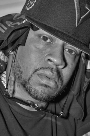 Close-up monochrome portrait of a man wearing a baseball cap, dark clothing, and sunglasses hanging around his neck by photographer Maggie Shannon.