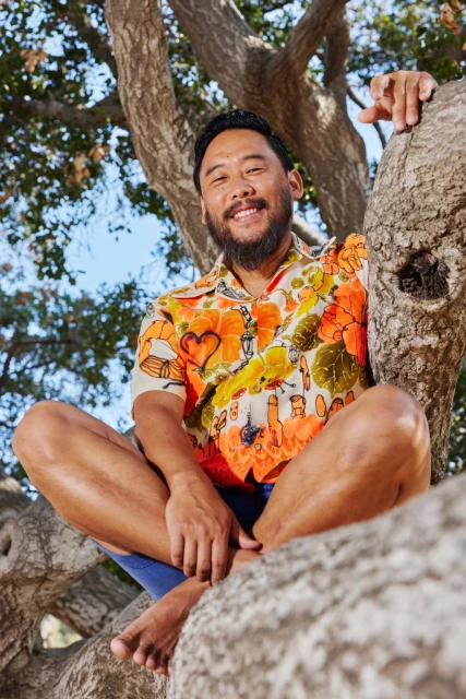 Portrait of a smiling Asian man sitting cross-legged on a tree branch wearing a vibrant, floral-patterned shirt in a natural setting.
