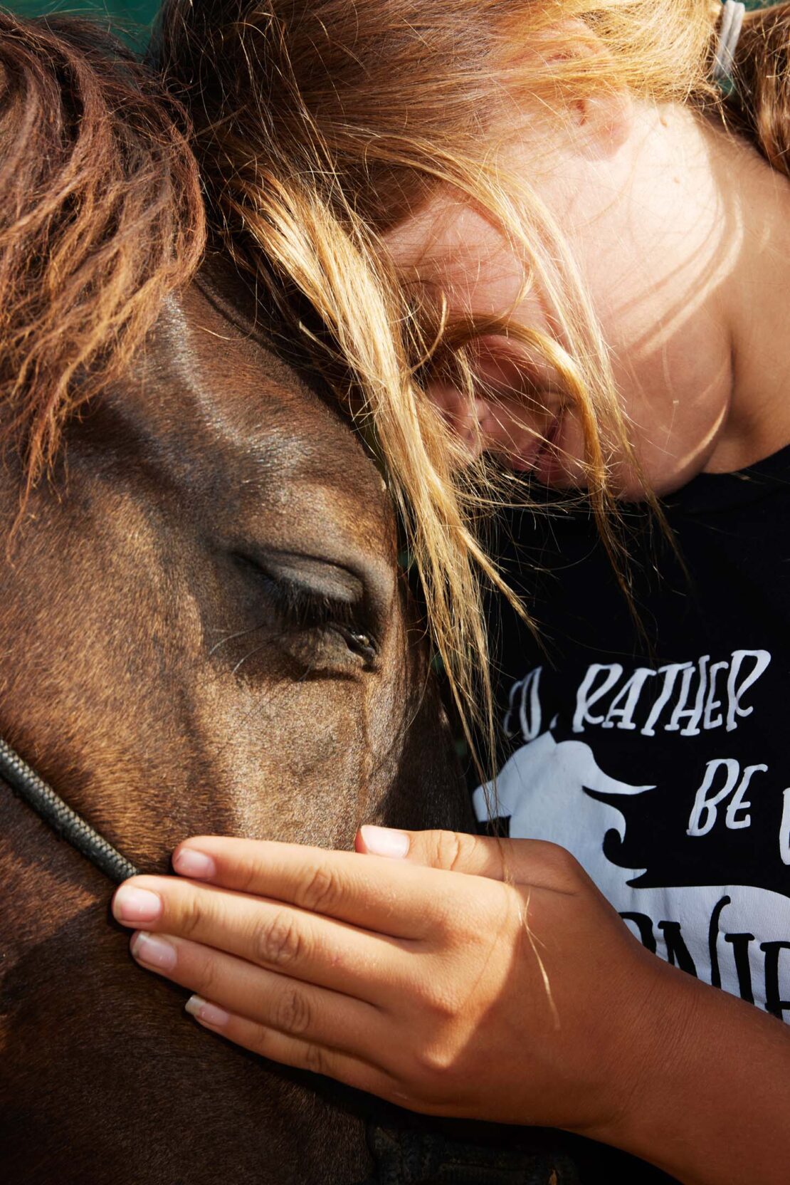 Close-up of a woman with long blonde hair embracing a brown horse. The horse's eye is visible, and the woman's face rests on the horse.