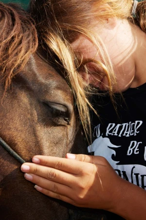 Close-up of a woman with long blonde hair embracing a brown horse. The horse's eye is visible, and the woman's face rests on the horse.