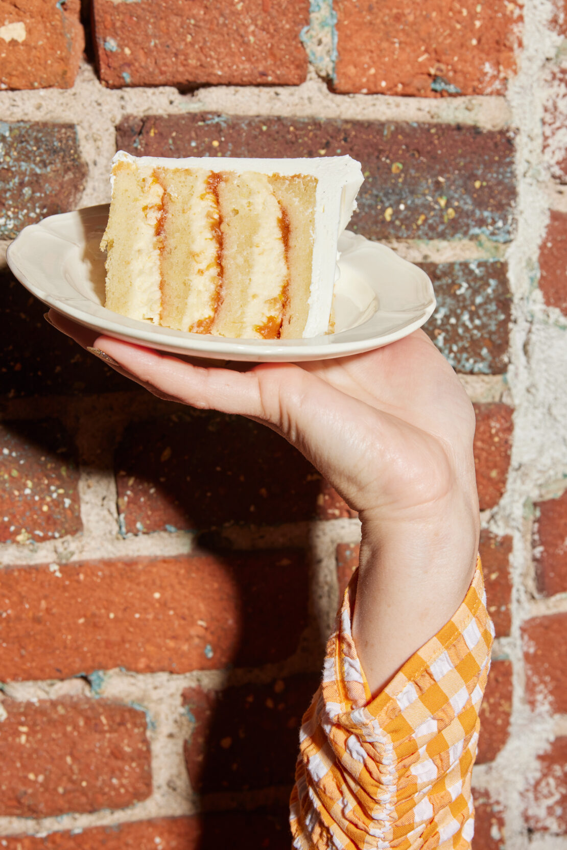 Hand holding a small white plate with a slice of layered cake against a red brick wall background.
