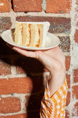 Hand holding a small white plate with a slice of layered cake against a red brick wall background.